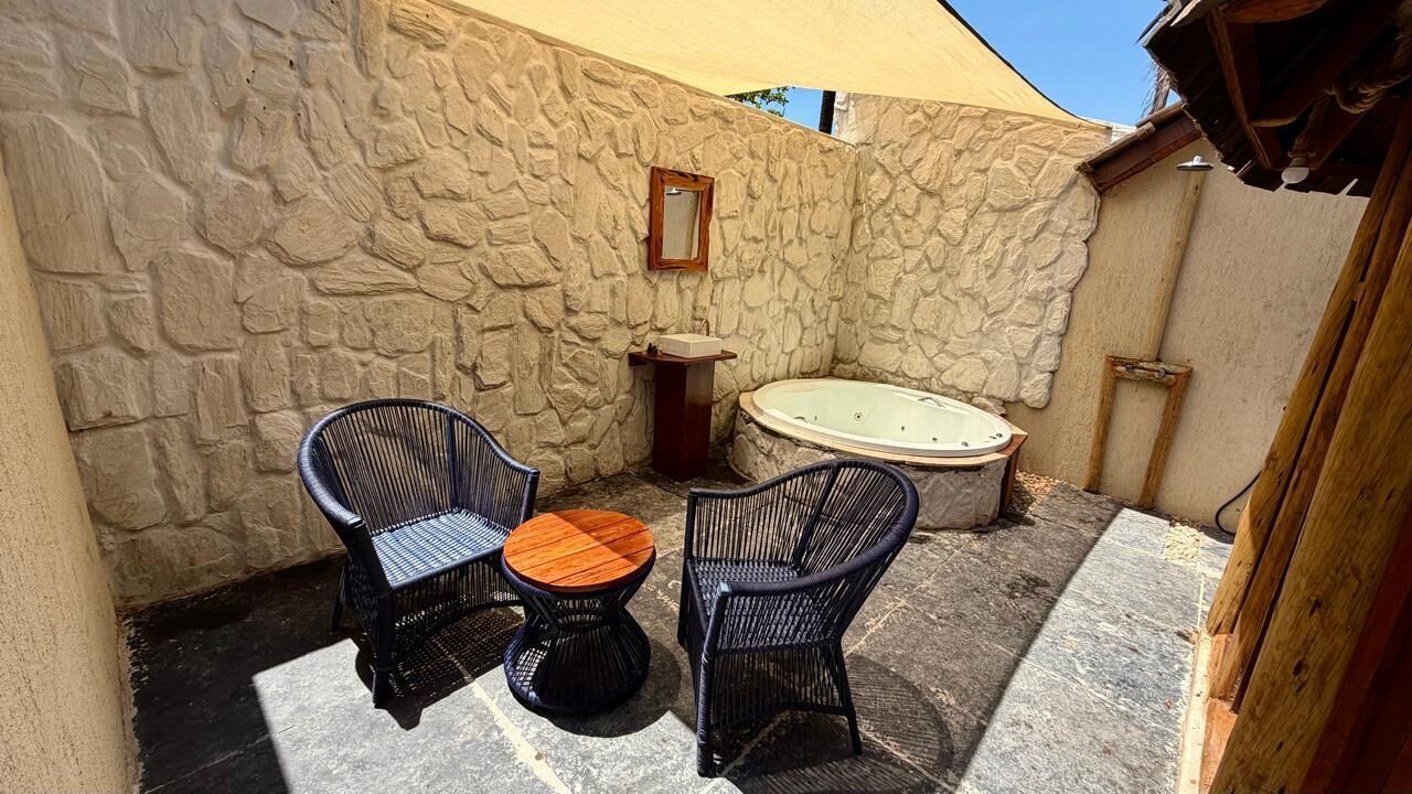 A stone-walled outdoor area with two wicker chairs and a small wooden table, a stone bench, and a hot tub.