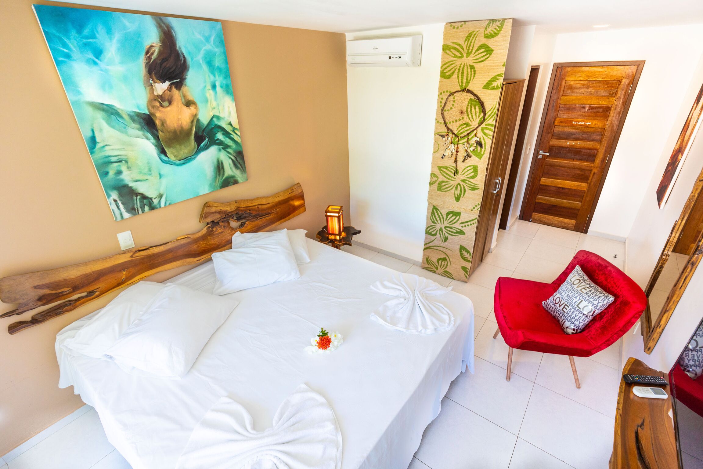Une chambre d’hôtel avec un grand lit, un fauteuil rouge, une tête de lit en bois, un tableau au mur et une porte avec un panneau en bois.