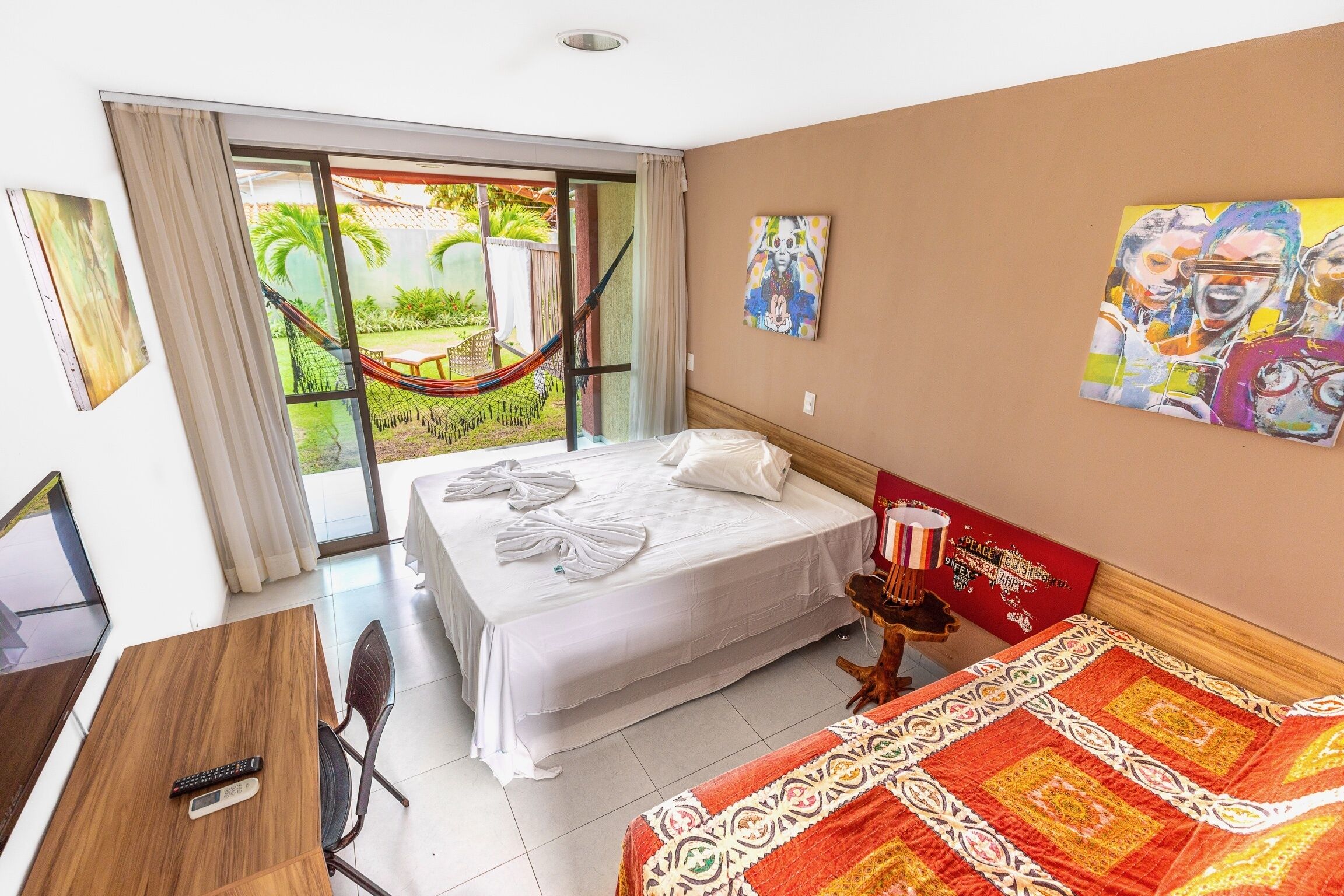 A hotel room with a bed, a desk, a chair, a window with a view of greenery, and a hammock.