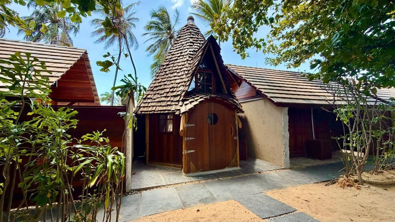 Um edifício rústico com porta de madeira e telhado inclinado, rodeado por vegetação tropical.