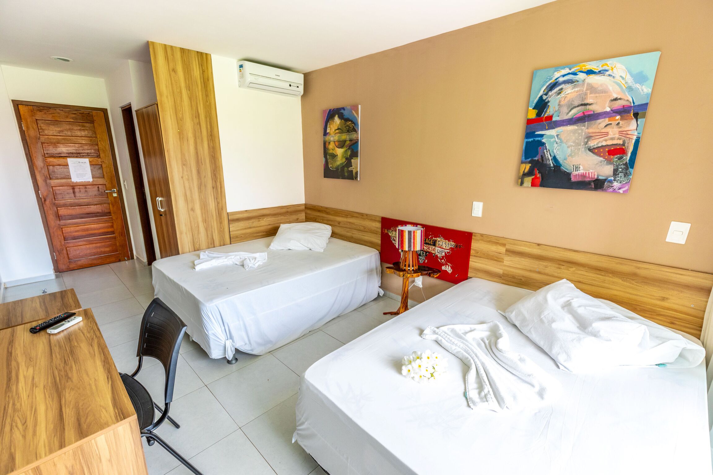 Une chambre d’hôtel avec deux lits simples, un bureau en bois avec une télécommande, une chaise et une petite table sur laquelle se trouve un livre rouge.
