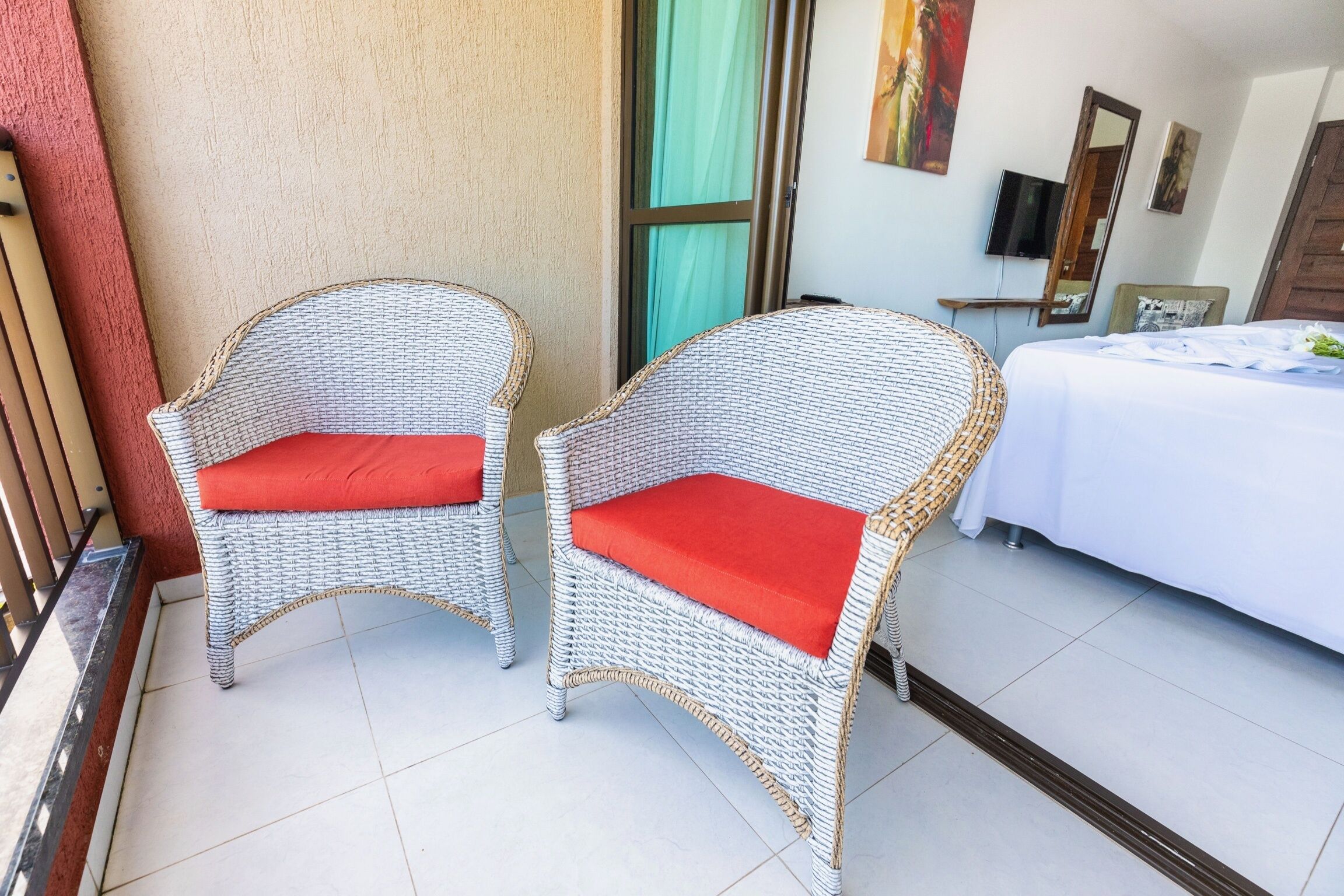 Una habitación con dos sillones de mimbre con cojines rojos, una cama con ropa de cama blanca y un televisor en la pared.