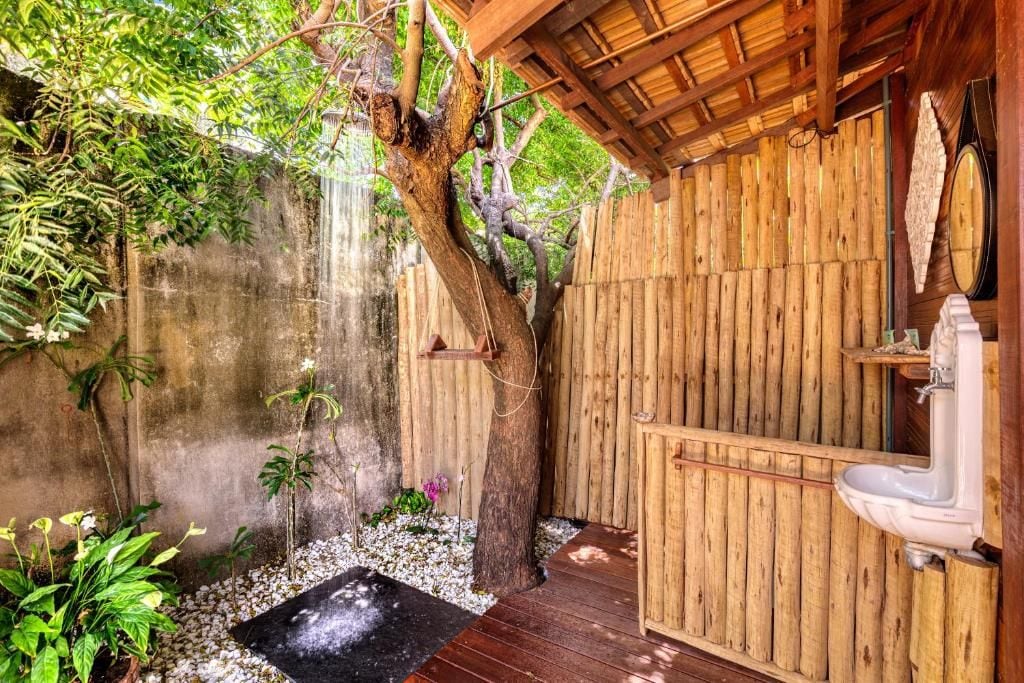 Un área de ducha exterior de madera con un árbol, una pequeña cascada y una terraza de madera.