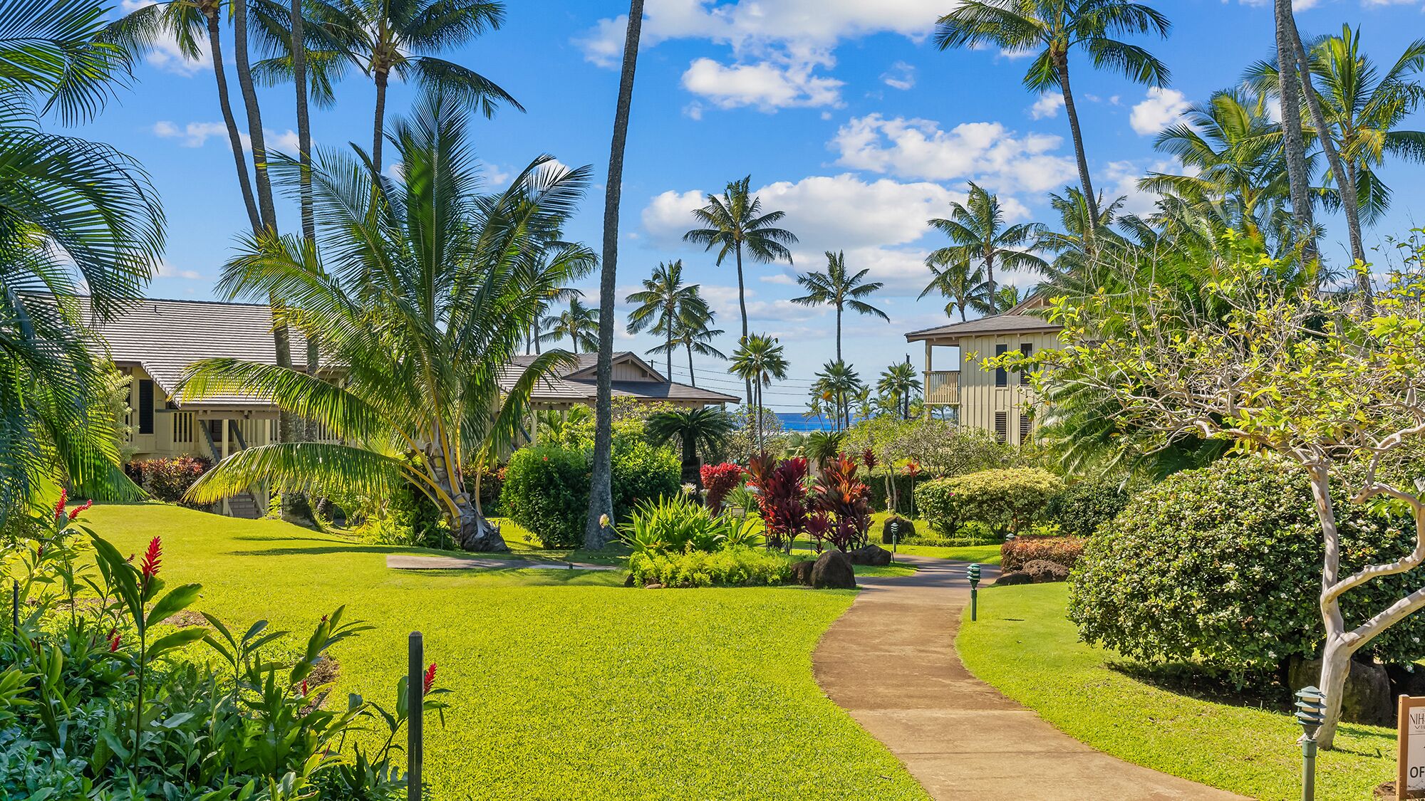 A tropical resort with a well-maintained lawn, palm trees, and a pathway leading to a building with a balcony overlooking the ocean.