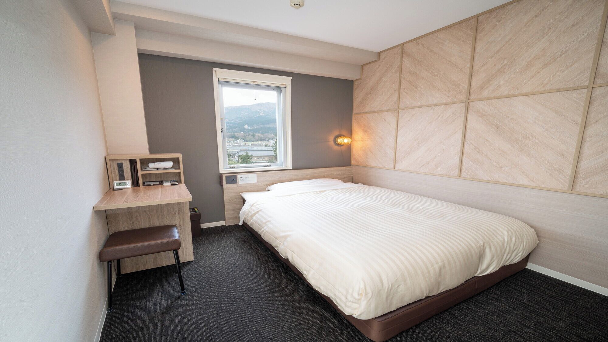 A hotel room with a bed, a desk, a chair, a window with a view, and a wall-mounted light.