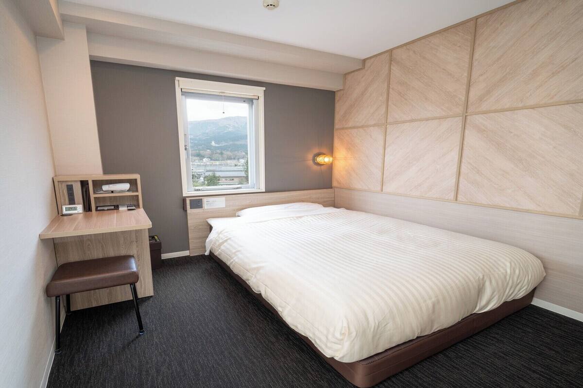 A hotel room with a bed, a desk, a chair, a window with a view, and a wall-mounted light.