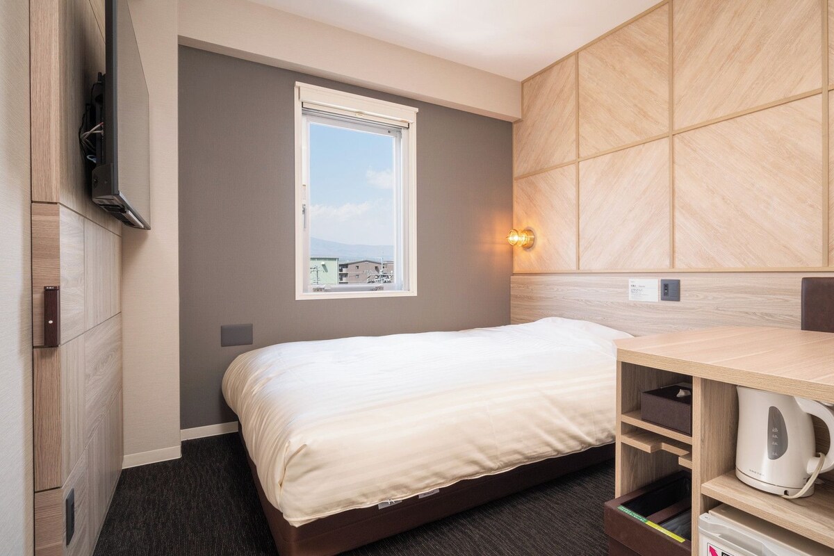 A hotel room with a bed, a desk with a kettle, a window with a view, and a wall-mounted TV.