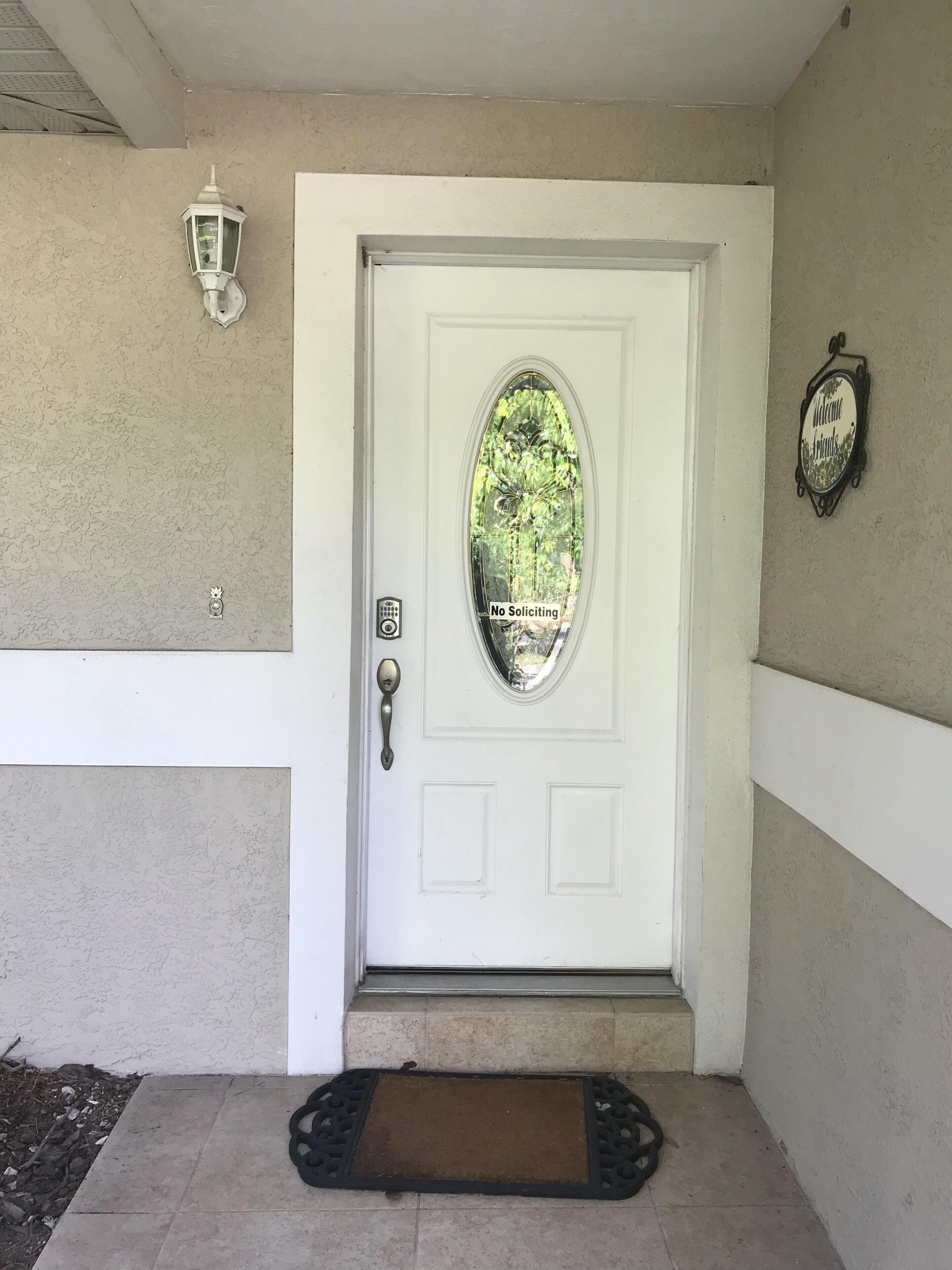 A white door with a frosted glass window, a doormat, a wall-mounted light, and a 'Welcome Friends' sign.