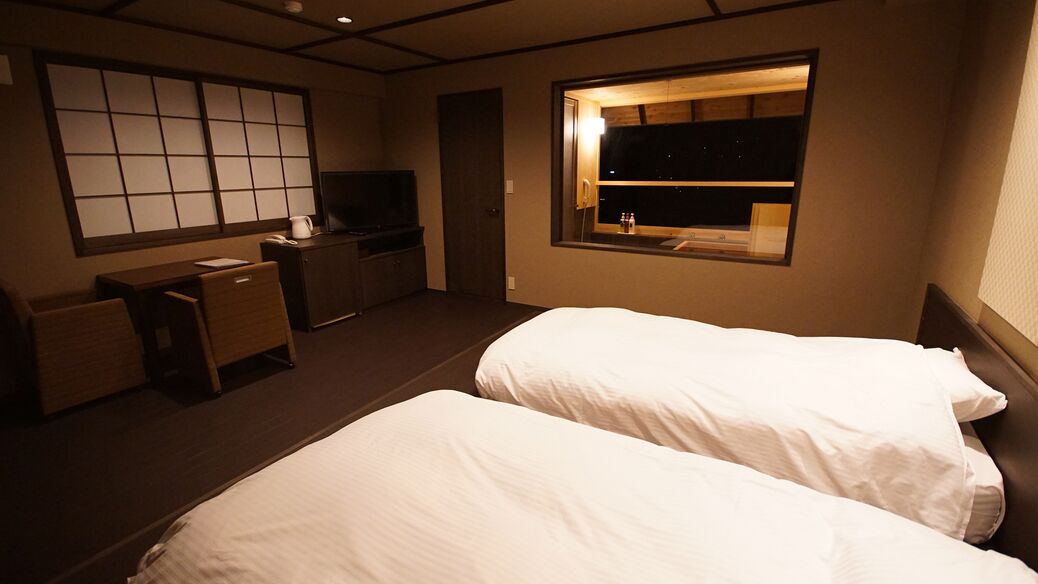 A hotel room with two beds, a desk, a television, and a window with a night view.