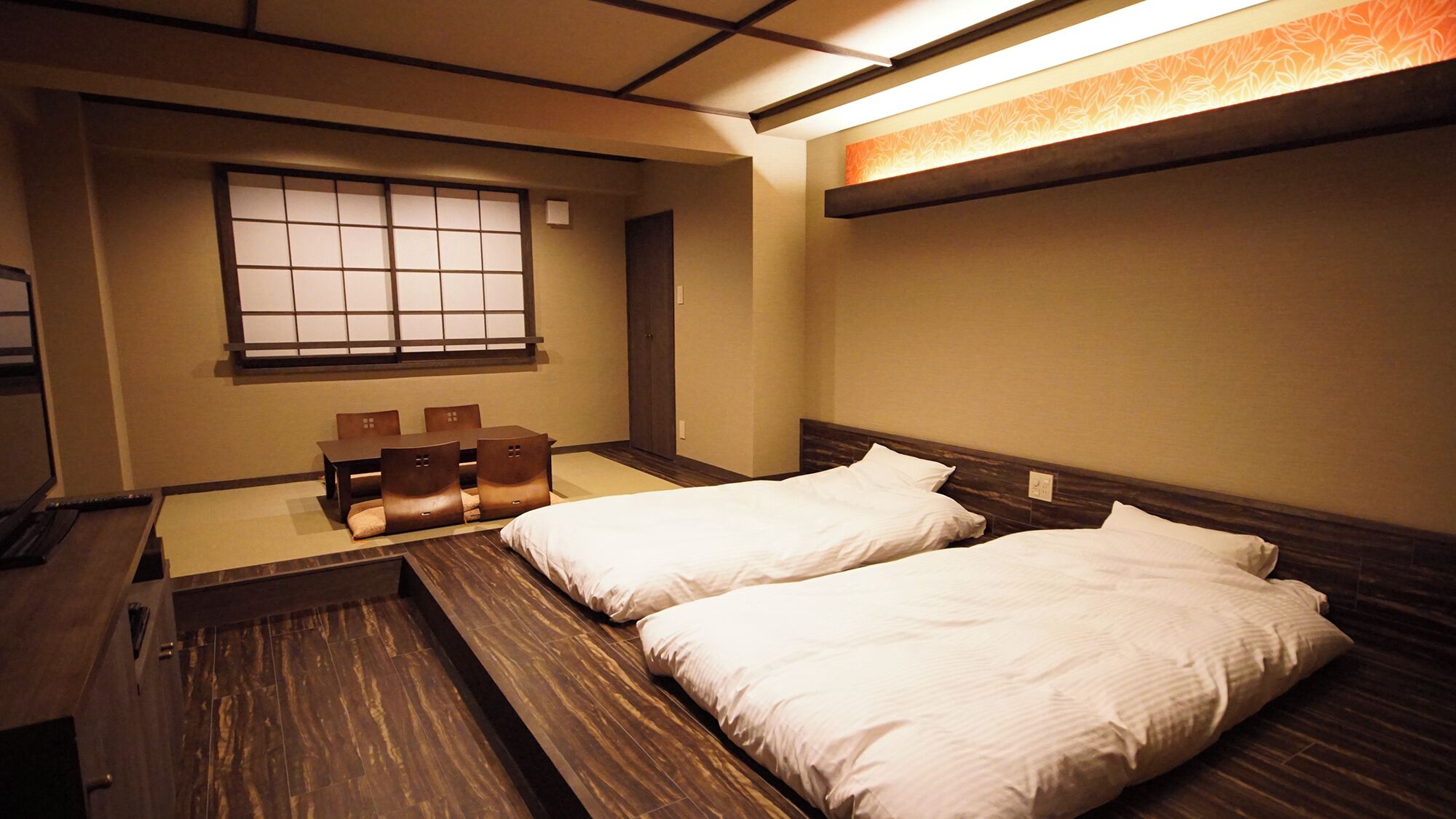 A room with two beds, a small table with chairs, a window, and a wall-mounted shelf.