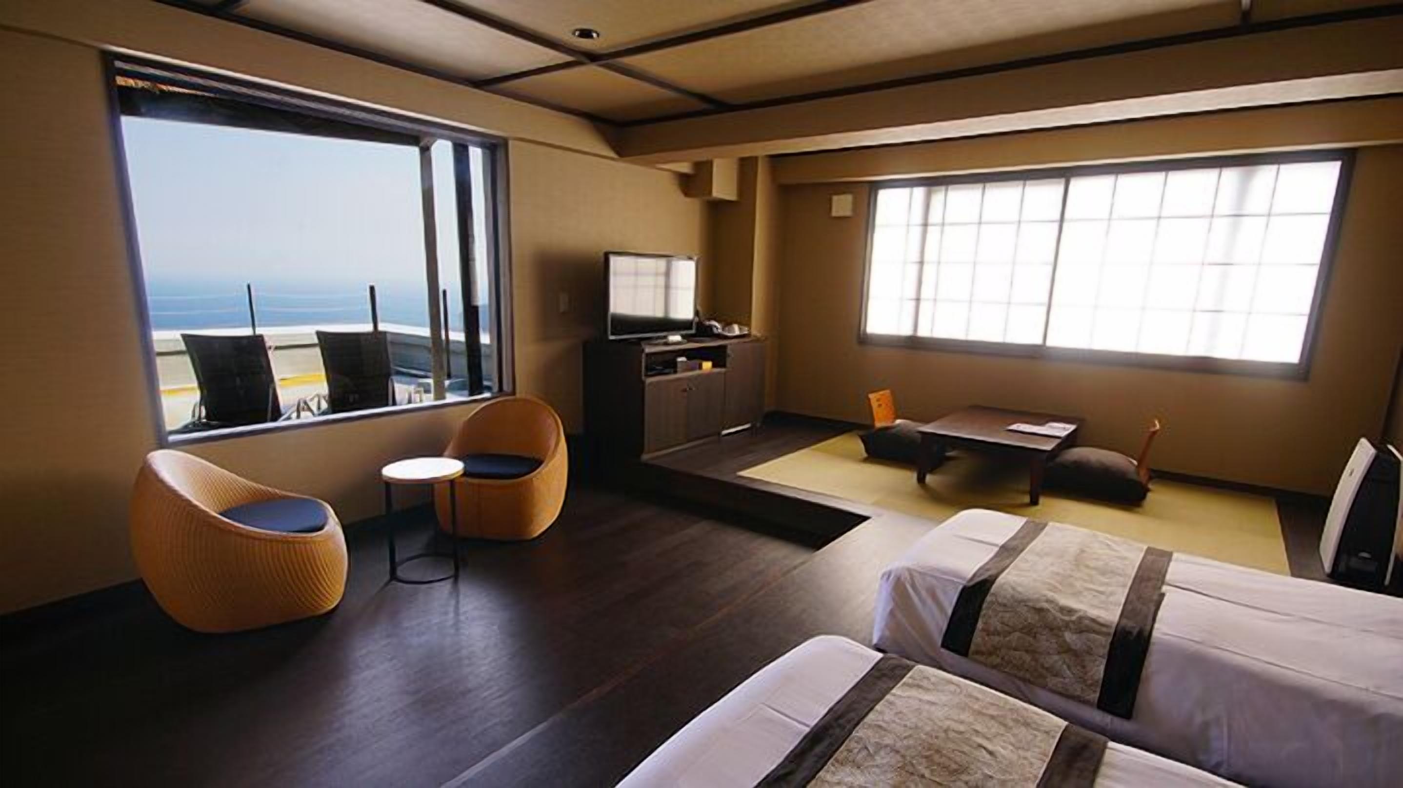 A hotel room with a large window, a television, a sofa, a small table, and two beds.