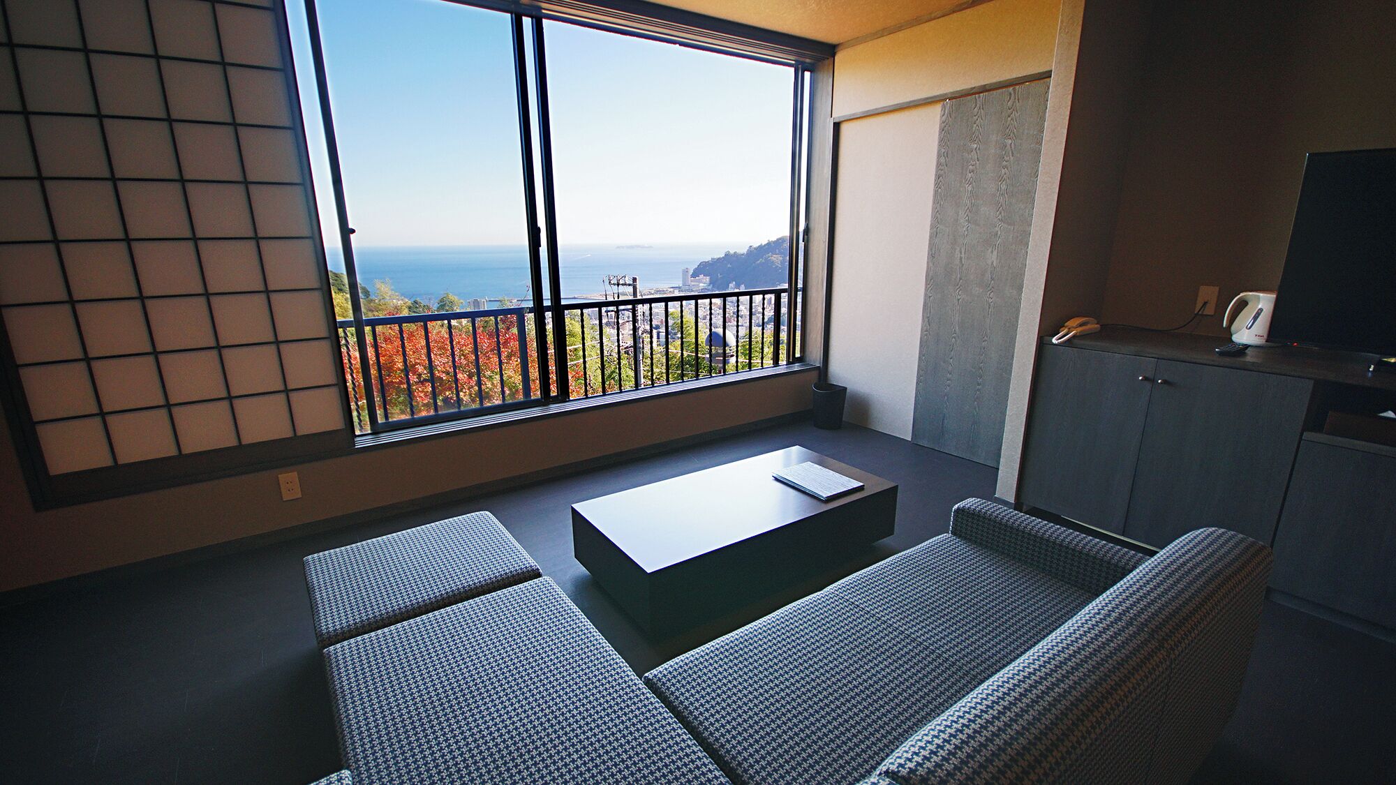 A modern living room with a large window, a sofa, a coffee table, and a small kitchen area.