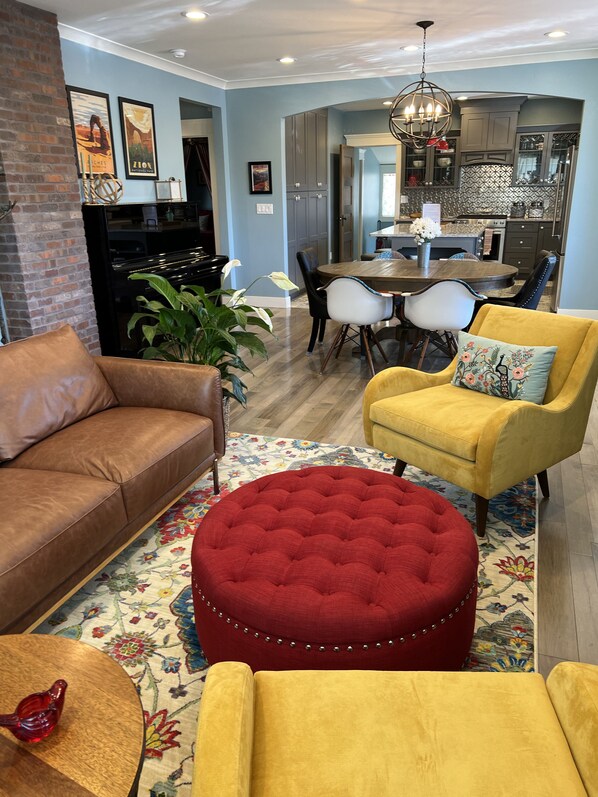 A living room with a brown leather sofa, a yellow armchair, and a red ottoman. There is a dining table with chairs, a kitchen in the background, and a chandelier. - Luxury Accomodations in updated 1920's bungalow in Downtown Provo (Provo)
