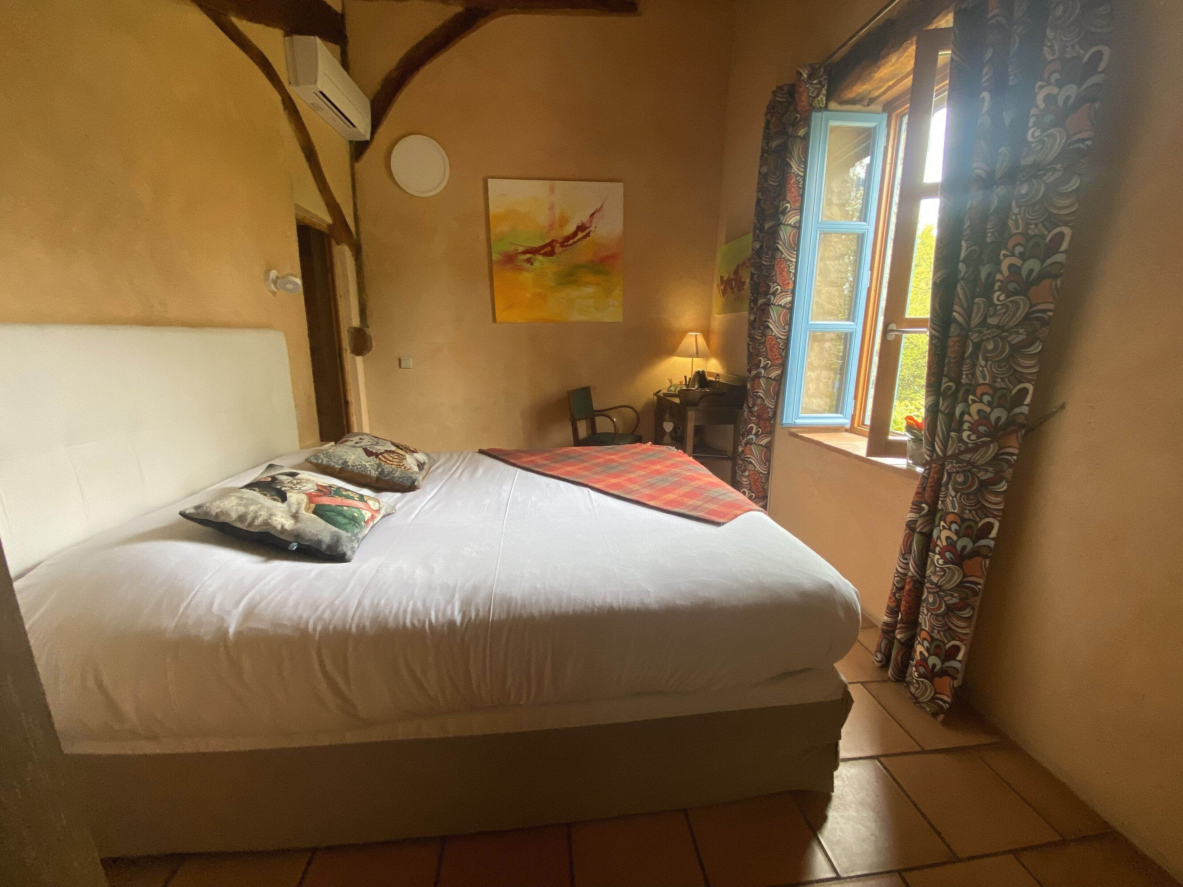 A bedroom with a bed, a desk, a chair, a window with curtains, and a wall-mounted air conditioner.