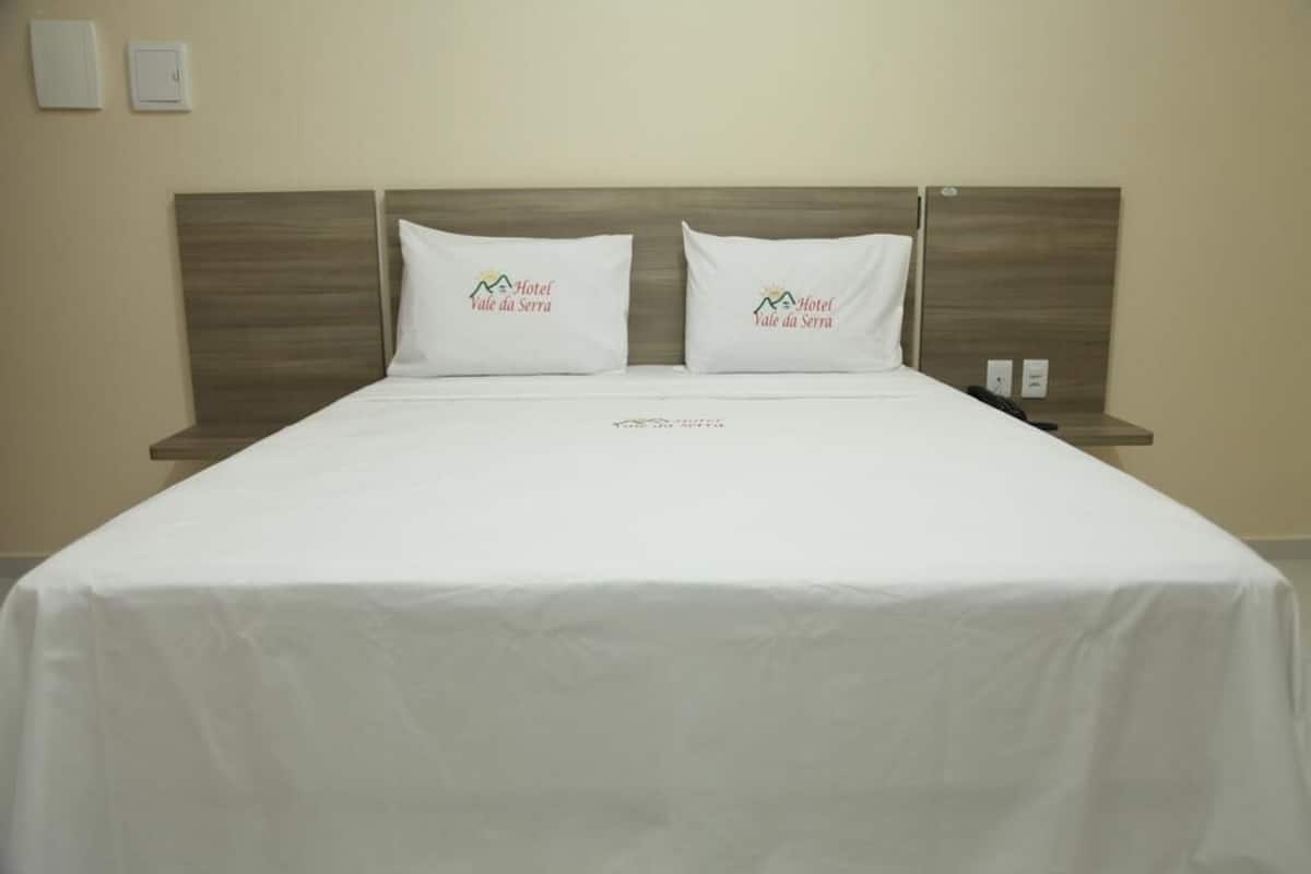 A hotel room with a large bed, two pillows with the hotel logo, a wooden headboard, and a nightstand with a phone.
