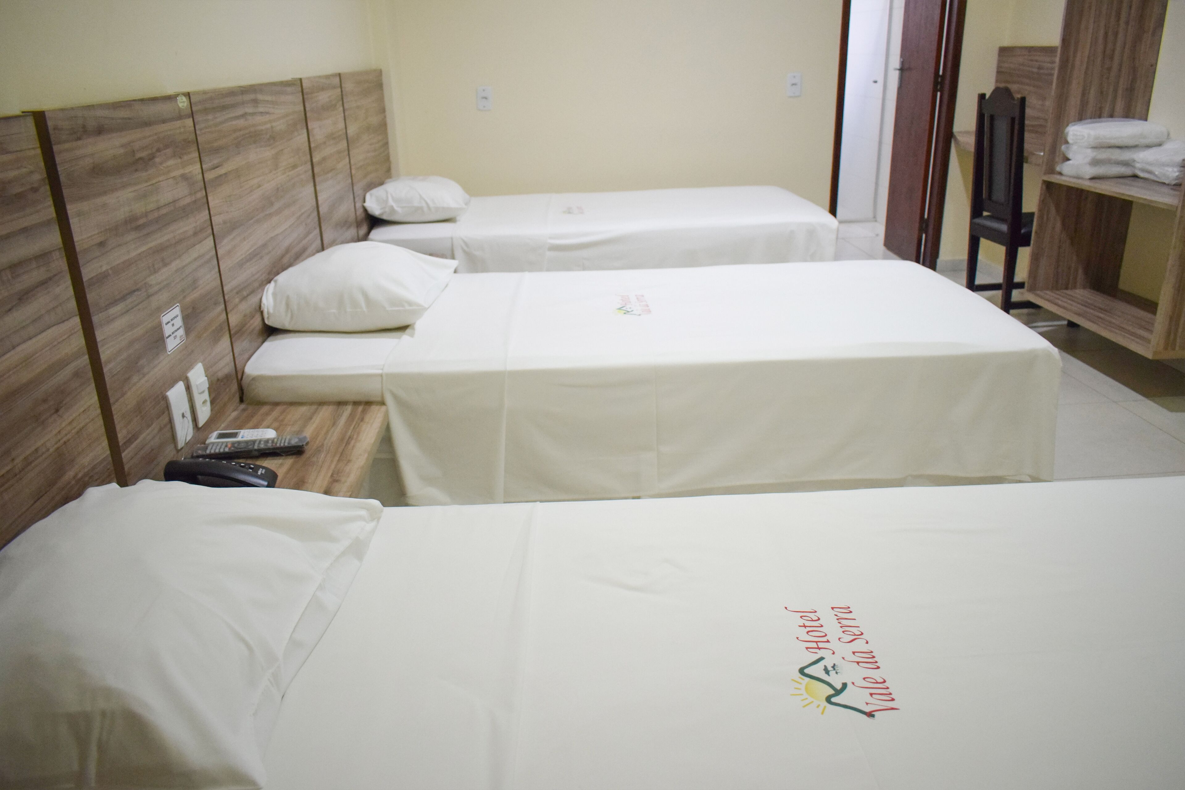 Habitación de hotel con dos camas individuales, cada una con almohada y manta. Se aprecia un cabecero de madera, una mesita con un control remoto y un estante con toallas dobladas.