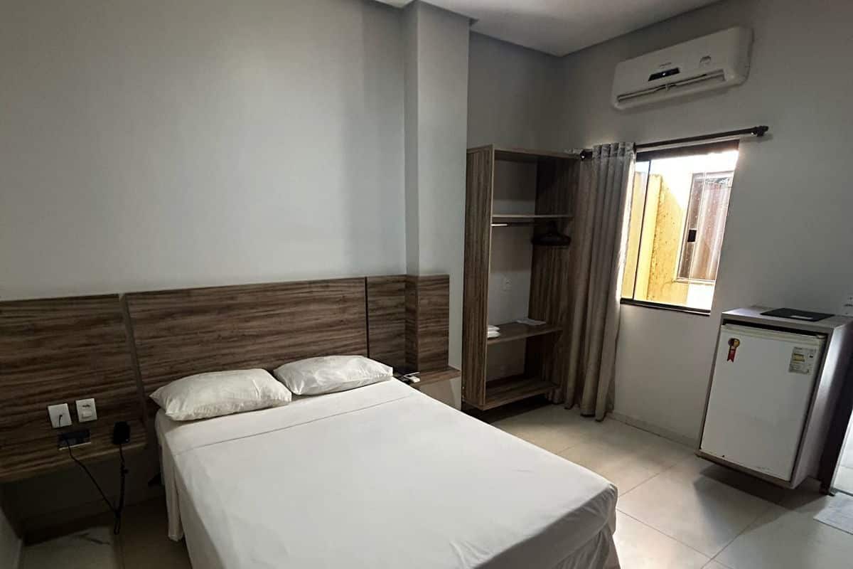 A hotel room with a bed, a wardrobe, a desk with a refrigerator, and a window with curtains.