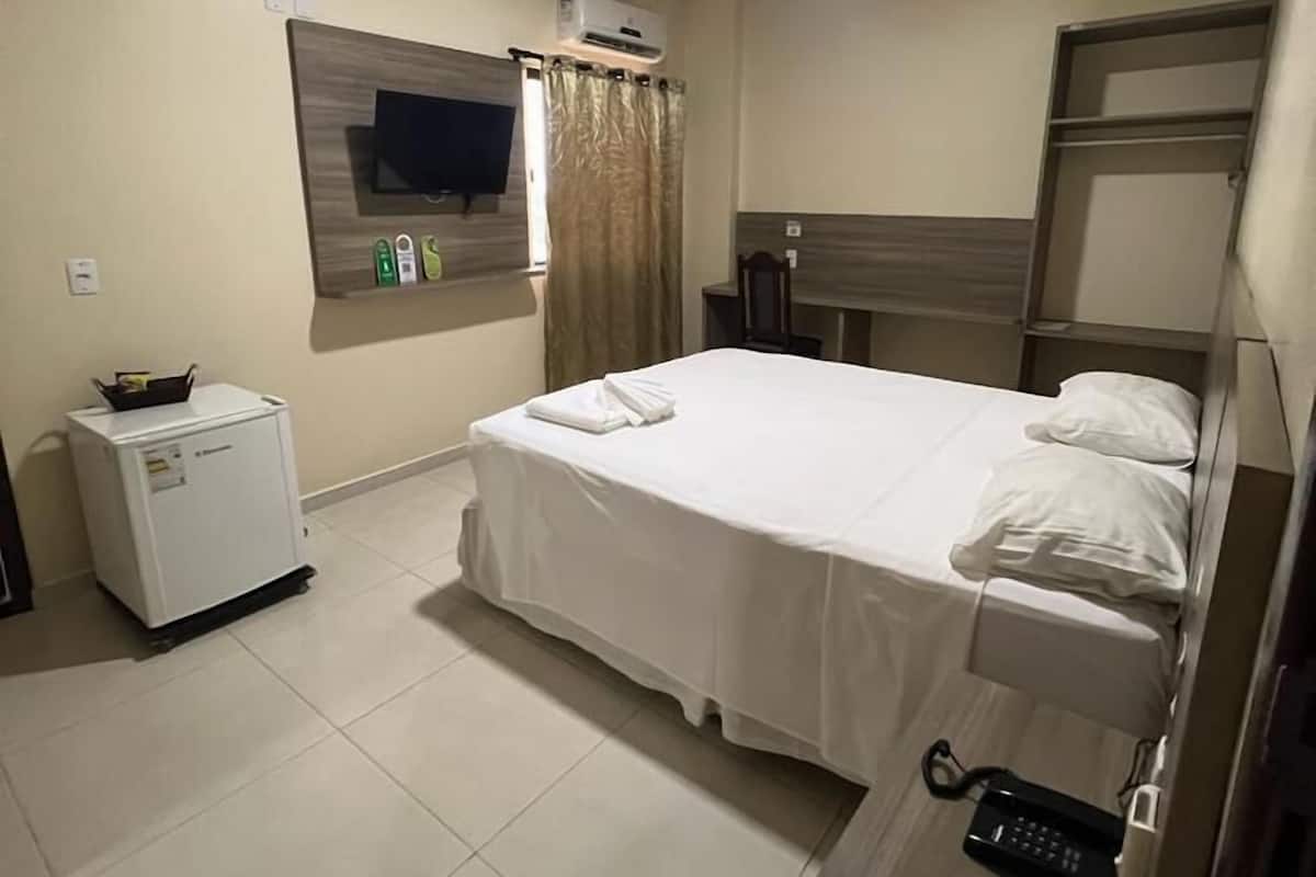 A hotel room with a bed, a TV mounted on the wall, a small refrigerator, and a bedside table with a remote control.