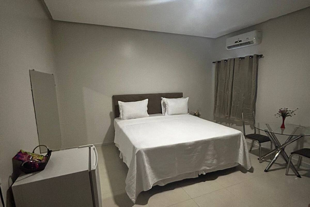 A hotel room with a bed, a small refrigerator, a table with chairs, and an air conditioning unit on the wall.
