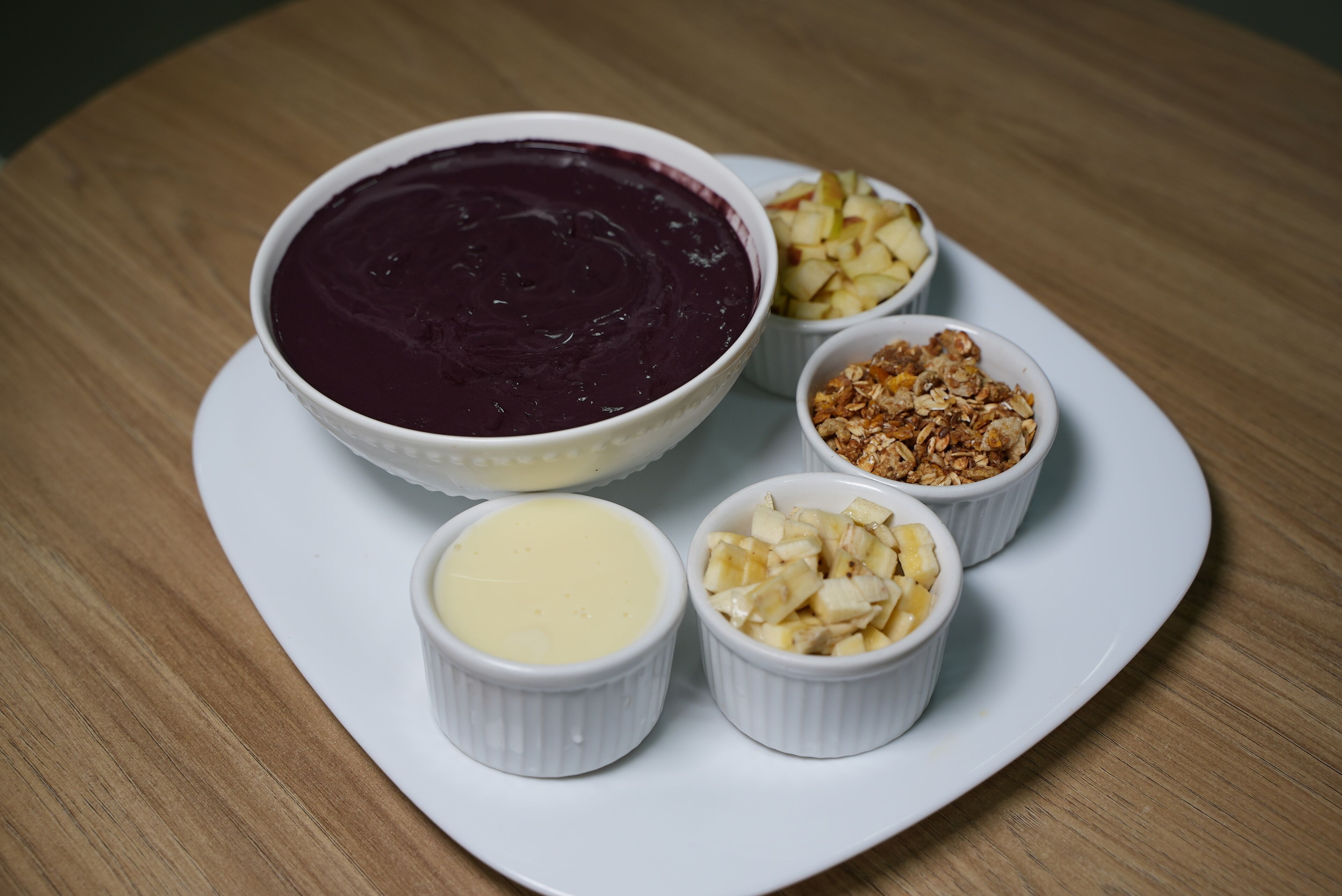 A white plate with a bowl of purple smoothie, a small bowl of yellow cream, and three small bowls containing different toppings.