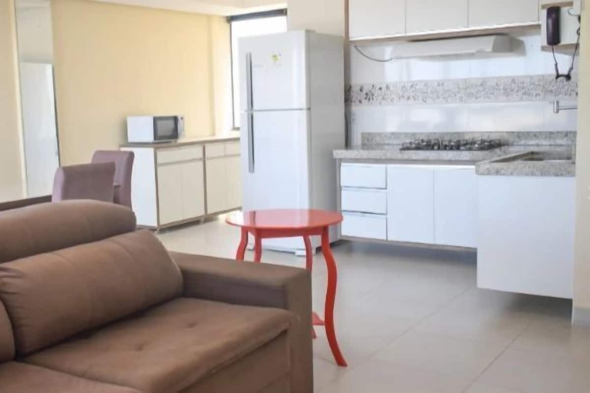 A living room with a brown sofa, a small red table, and a kitchen area with white cabinets and a microwave.