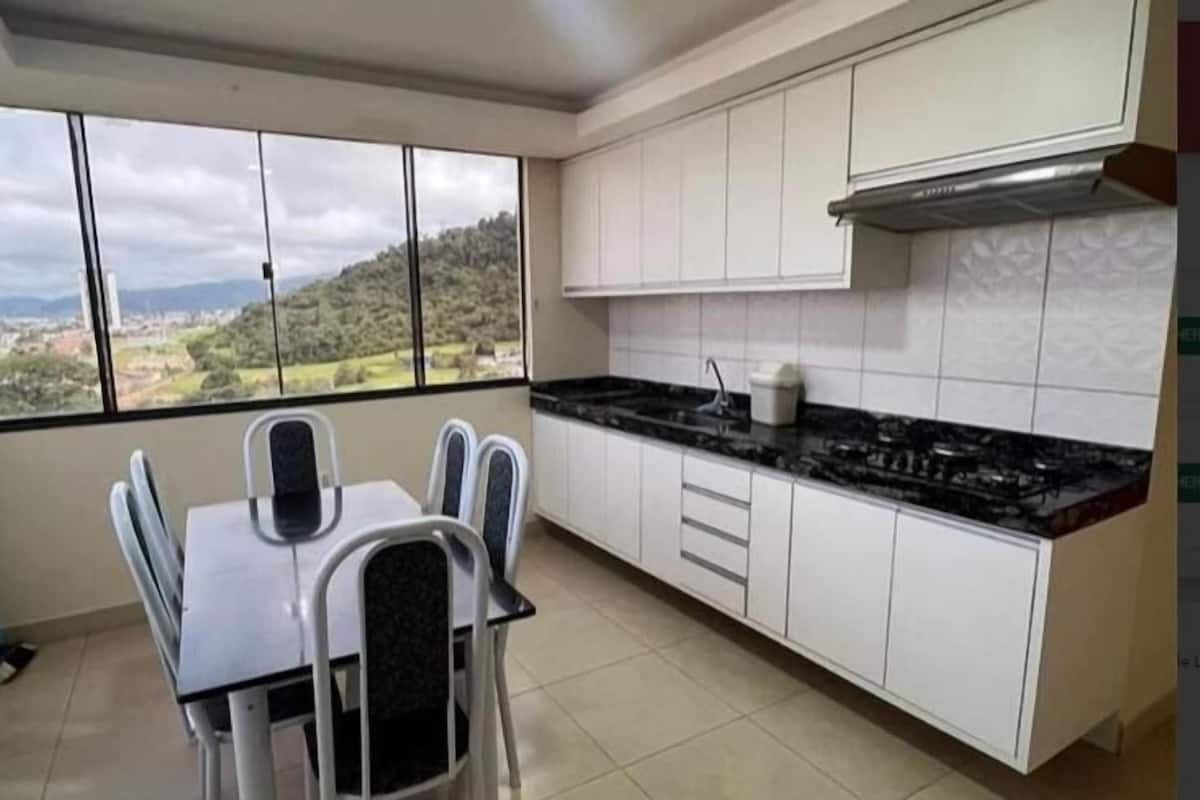 A kitchen with a dining table and chairs, white cabinets, a black countertop, and a view of a green landscape through large windows.