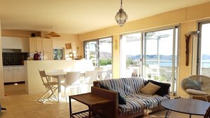 A spacious living area with a sofa, a coffee table, and a dining table. There is a kitchen area with cabinets and a view of the outdoors. - Holiday house facing the sea (Trébeurden)