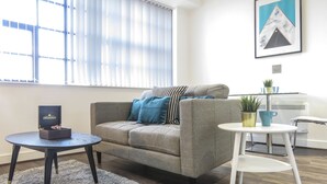 A modern living room with a grey sofa, a small round table, and a white side table. There is a framed picture on the wall and a window with blinds. - Tudors eSuites Birmingham Apartments Jewellery Quarter (Birmingham)