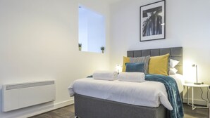 A modern bedroom with a grey upholstered bed, white linens, and a variety of pillows. There is a bedside table with a lamp, a radiator, and a window with a view of the sky. - Tudors eSuites Birmingham Apartments Jewellery Quarter (Birmingham)