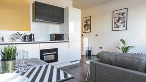 A modern kitchen with white cabinets, a black microwave, and a built-in oven. There is a grey sofa and a striped table in the living area. - Tudors eSuites Birmingham Apartments Jewellery Quarter (Birmingham)