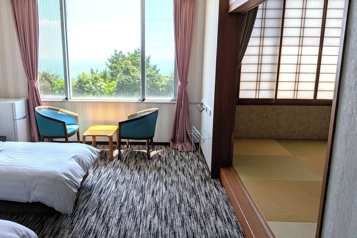 Habitación de hotel con cama, dos sillas, una mesita y vistas a la vegetación por la ventana.