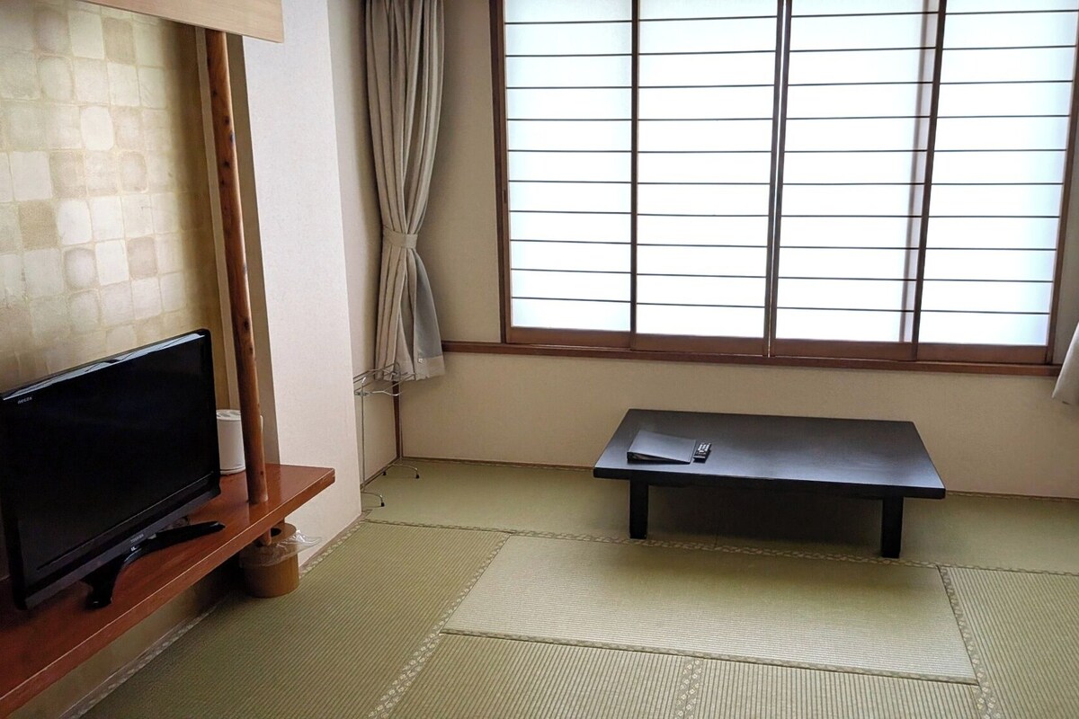 Una habitación con esteras de tatami, un televisor, una mesita y una ventana con cortinas.
