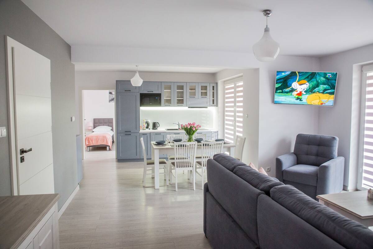 Ein modernes Wohnzimmer mit grauem Sofa, grauem Sessel und einem Essbereich mit weißem Tisch und Stühlen.