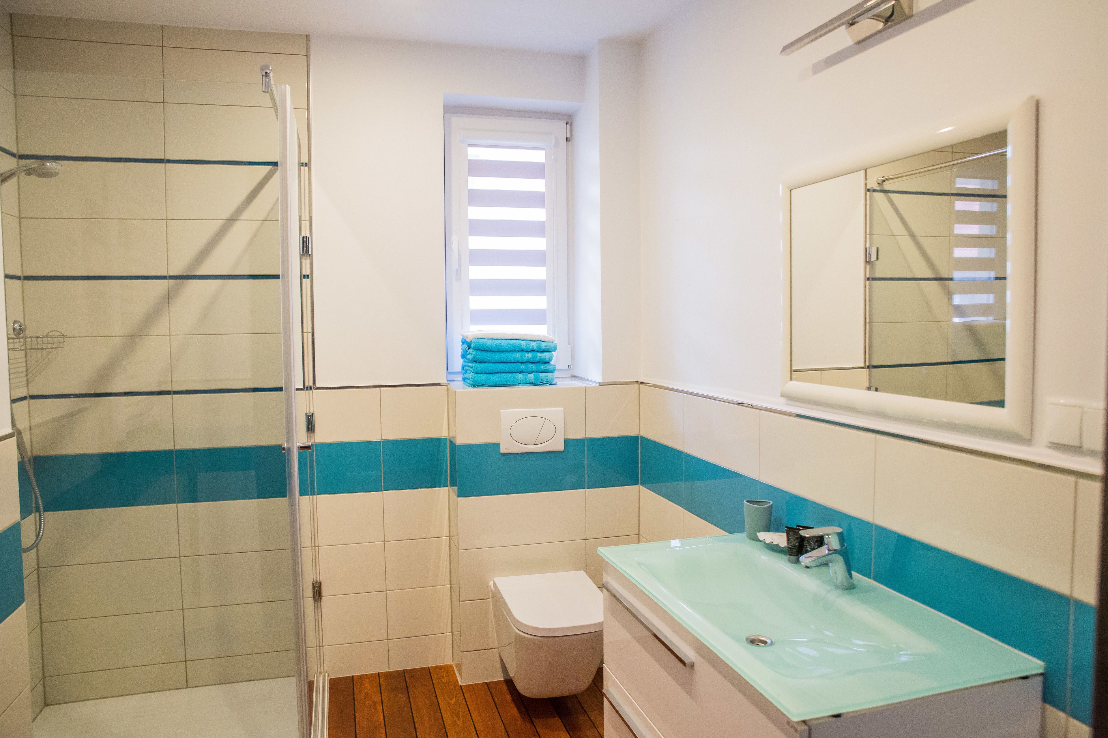 A bathroom with a glass shower enclosure, a toilet, a sink with a glass basin, and a window with blinds.