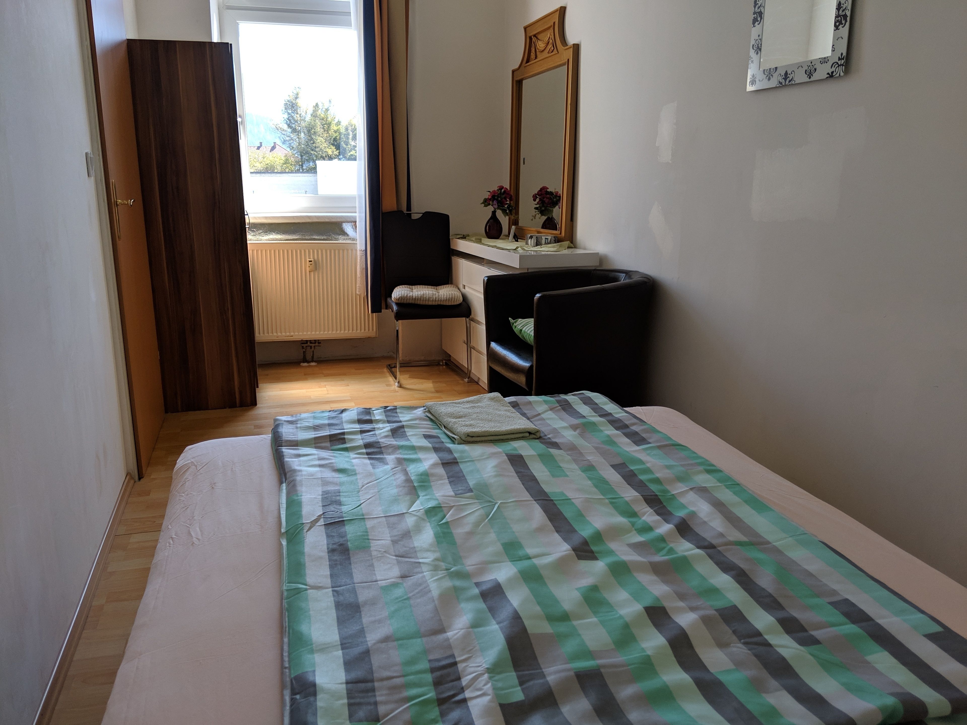 A bedroom with a bed, a chair, a dresser, a mirror, and a window with a view of the outside.