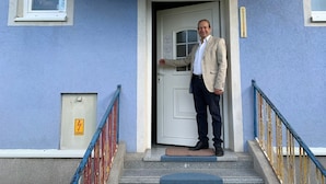 Un hombre está de pie frente a la entrada de un edificio con puerta blanca, paredes azules y barandilla azul.
