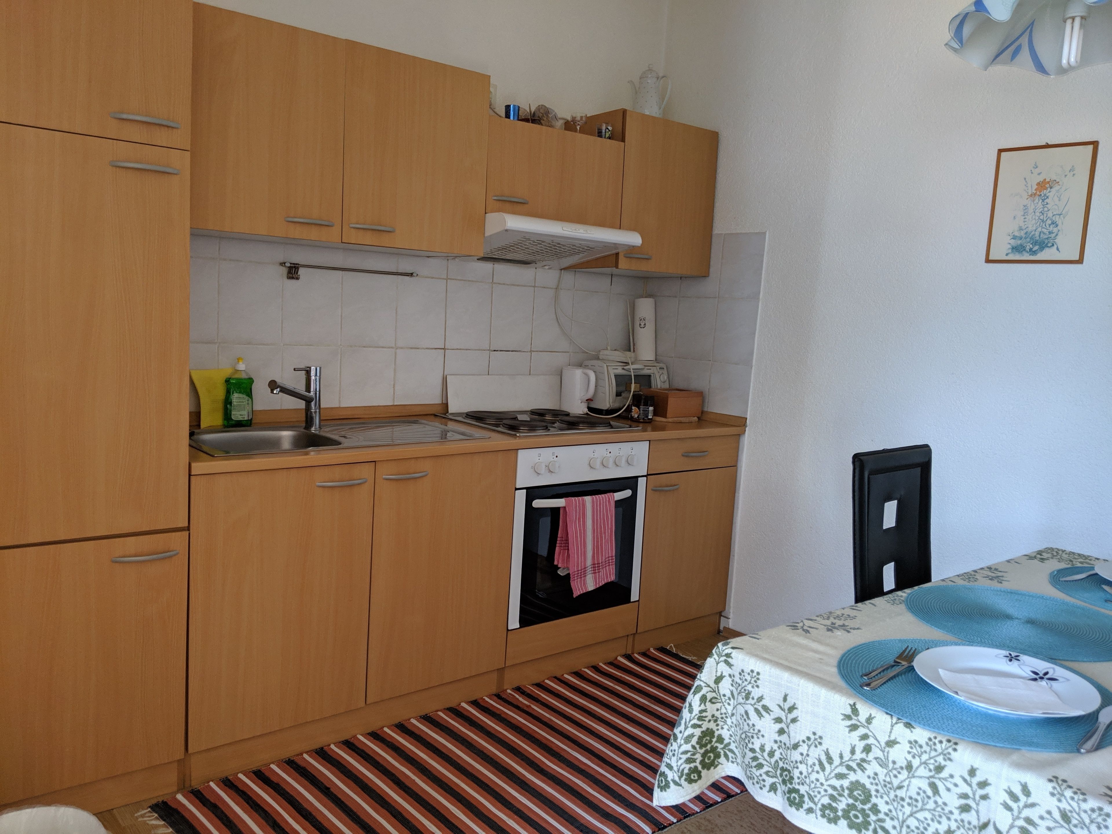 A small kitchen with wooden cabinets, a stove, a sink, and a dining area with a table set for two.