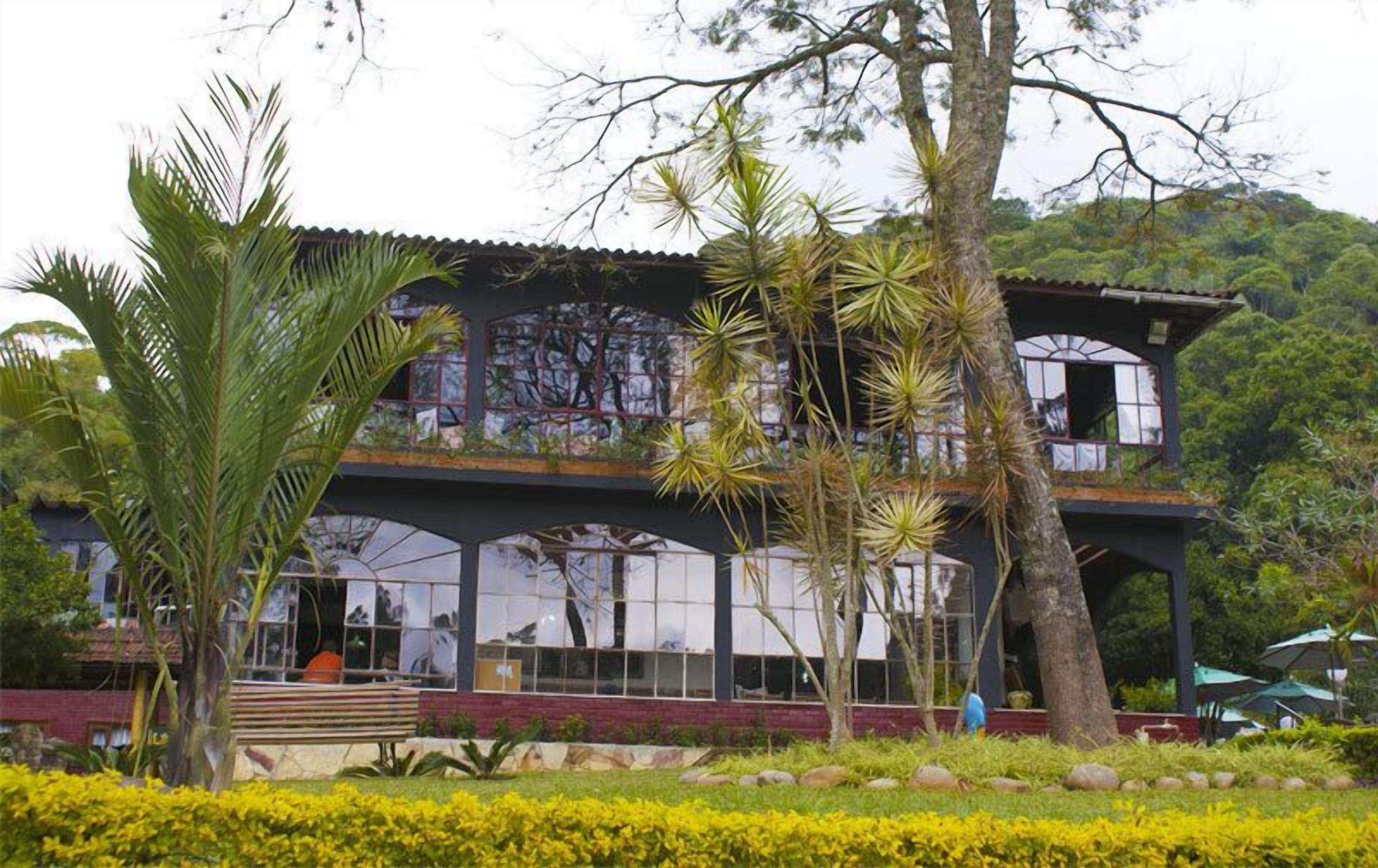 Um edifício de dois andares com janelas amplas e uma varanda, cercado por plantas tropicais e um jardim paisagístico.