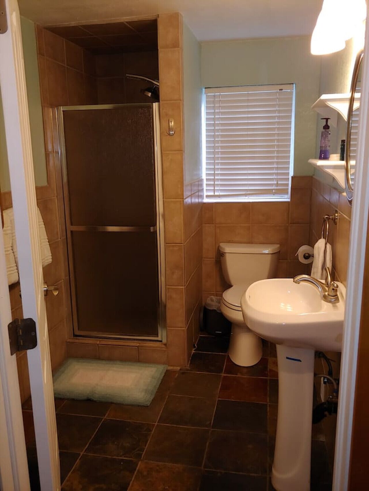 A bathroom with a shower, toilet, sink, and a window with blinds.