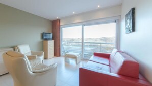 Een moderne woonkamer met een rode bank, witte fauteuils, een televisie op een kast en een grote schuifpui naar een balkon.