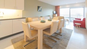 Een moderne keuken met witte kasten, een eettafel met zes stoelen, en een rode bank in een lichte woonruimte met grote ramen.
