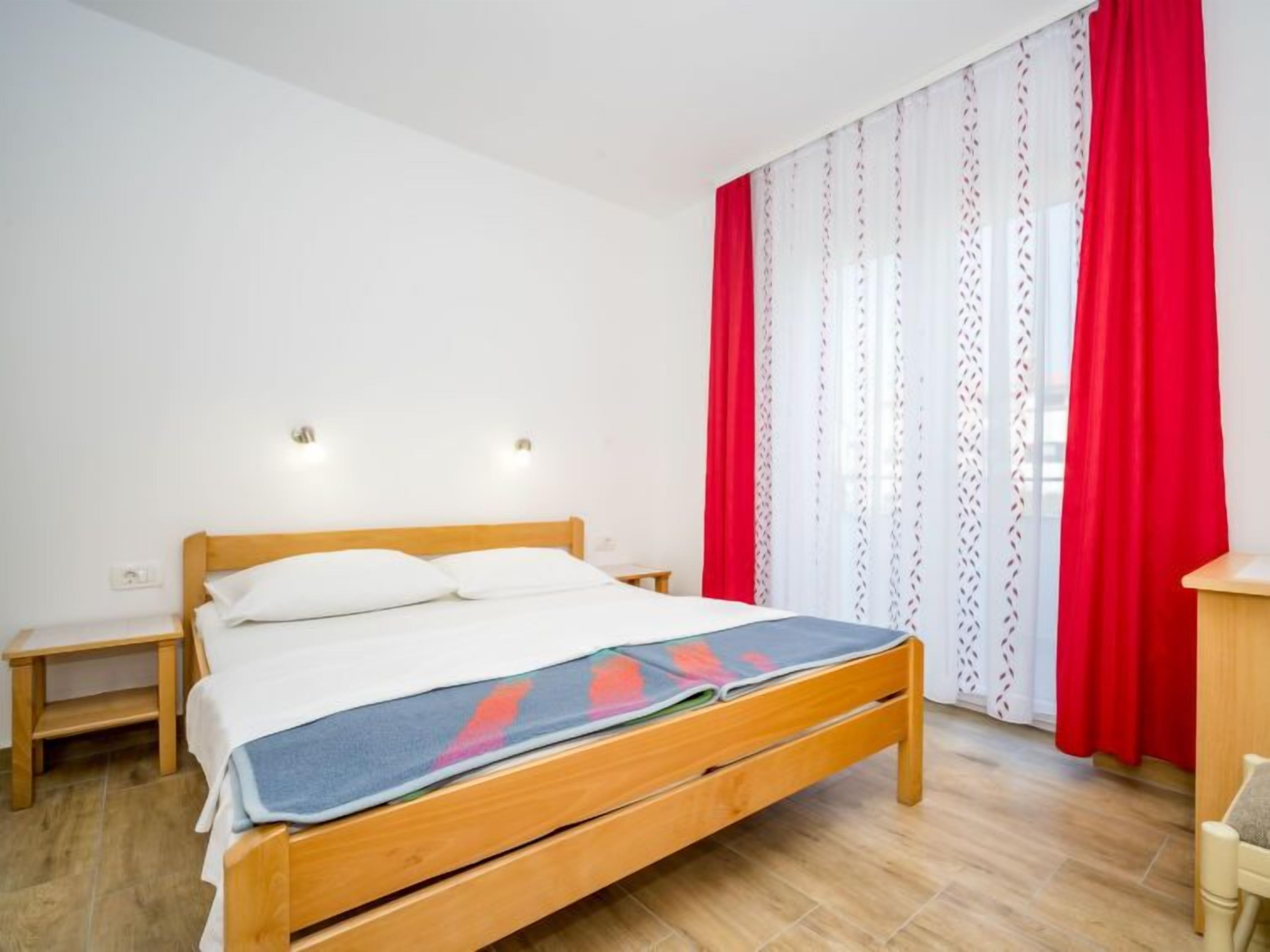 A bedroom with a wooden bed, white bedding, a nightstand, a small table, and a window with red curtains.