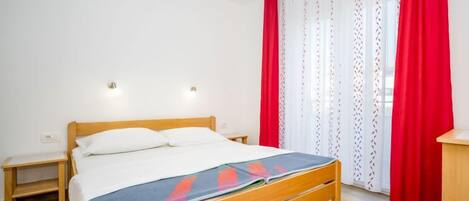 A bedroom with a wooden bed, white bedding, a nightstand, a small table, and a window with red curtains.