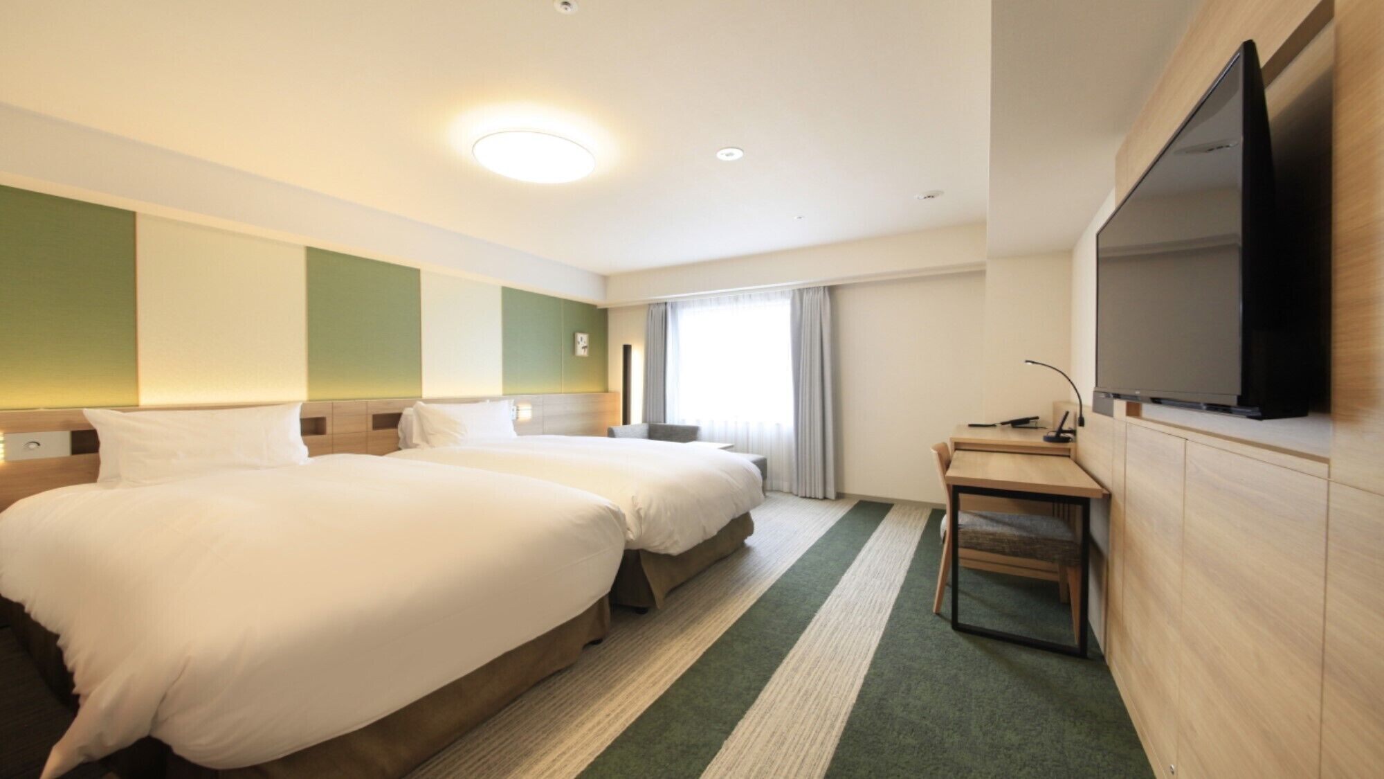 A hotel room with two beds, a desk, a chair, a flat-screen TV, and a large window.