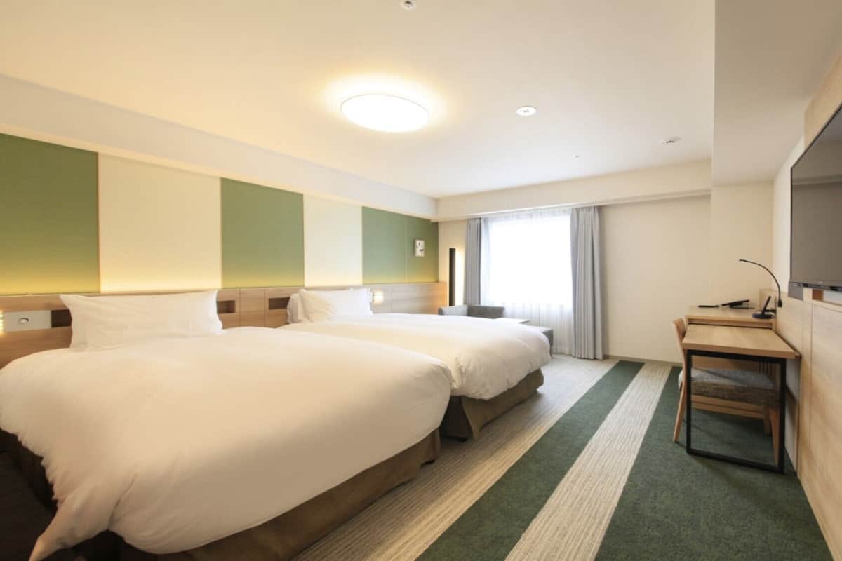 Quarto de hotel com duas camas, uma escrivaninha, uma cadeira, televisão de tela plana e uma janela grande.