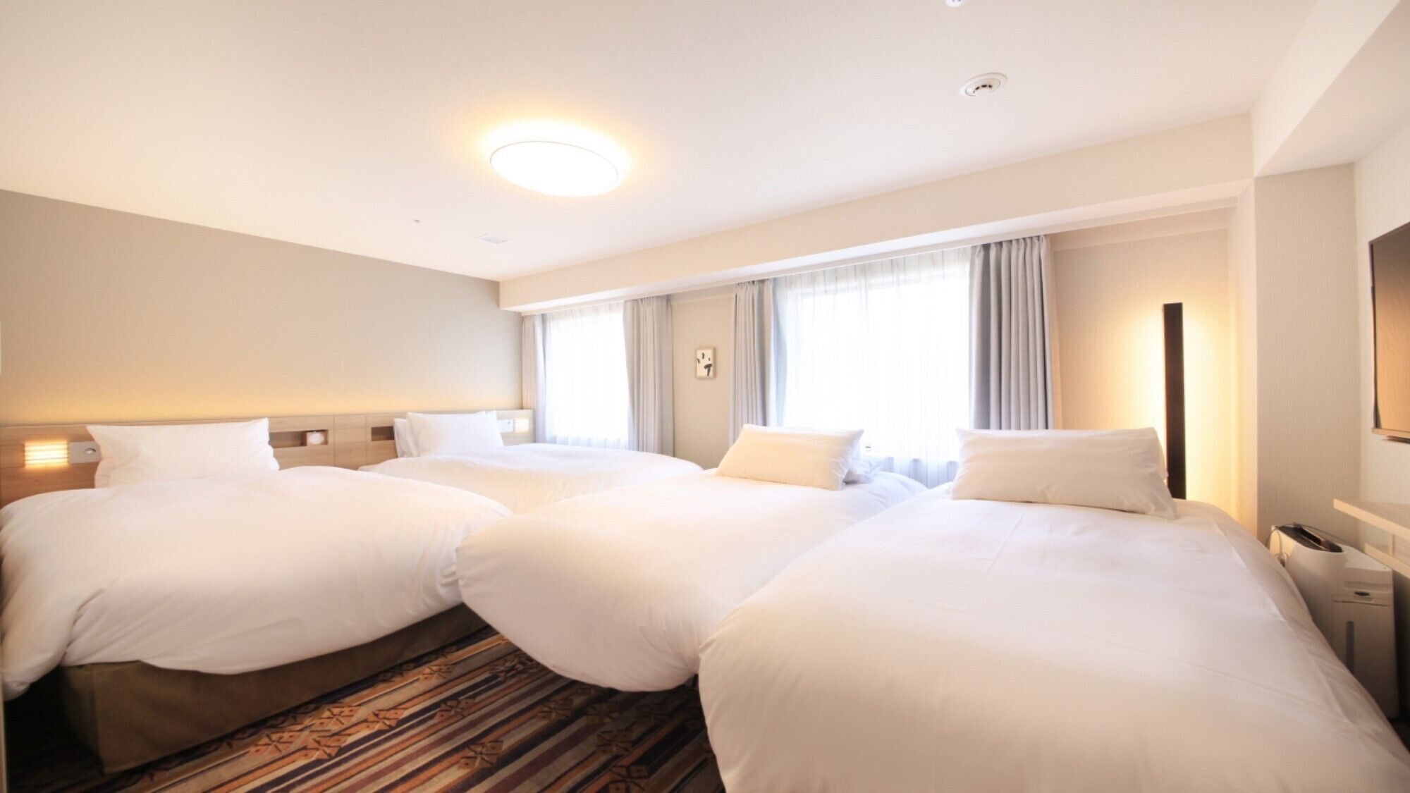 A hotel room with two beds, a large window with curtains, a flat-screen TV, and a bedside table with a lamp.