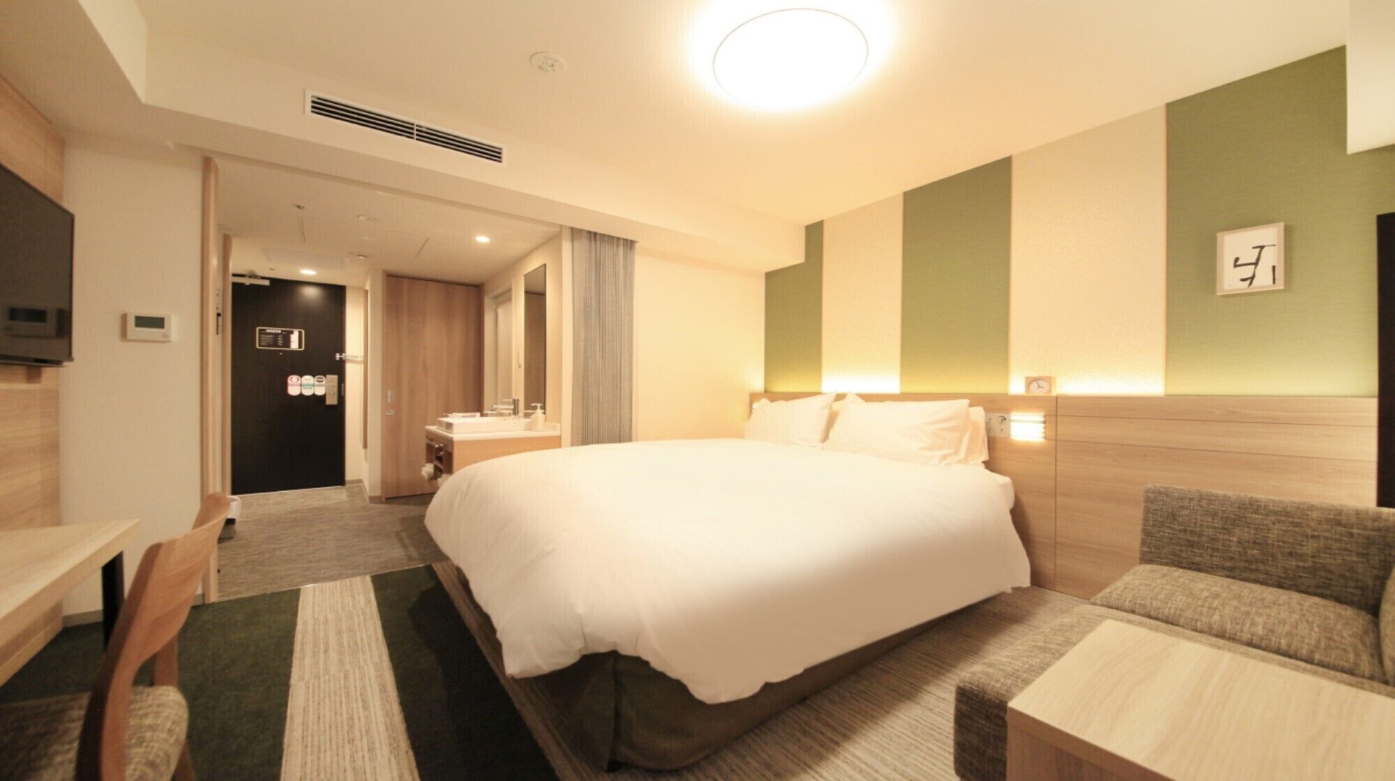 A hotel room with a bed, a desk, a chair, a mirror, a bedside table, and a wall clock.