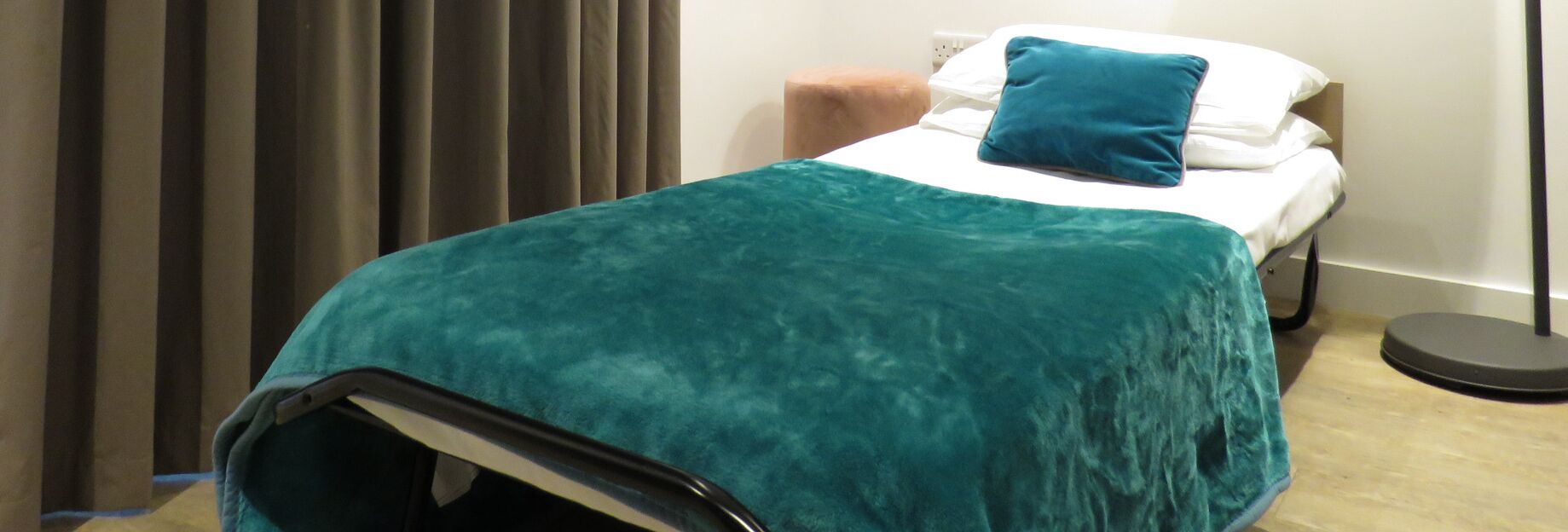A single bed with a teal blanket and a blue pillow in a room with a wooden floor and a curtain.