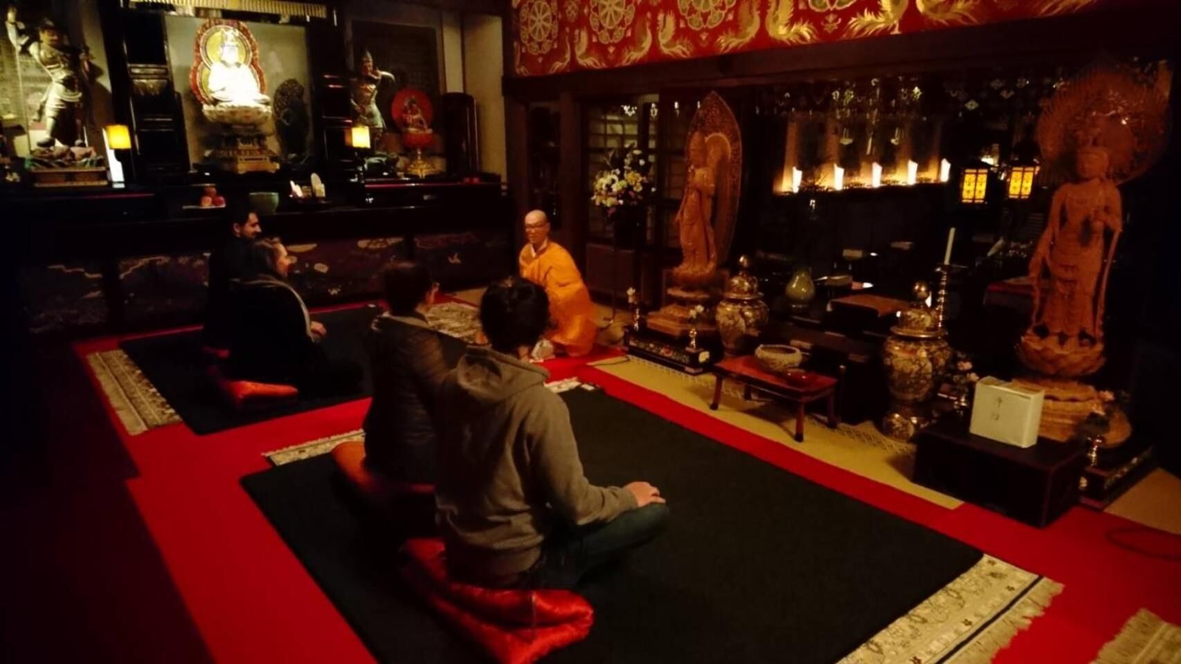A room with a red carpet, a seated person in orange robes, and various statues and decorations.