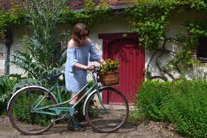 A woman standing next to a vintage bicycle with a basket, in front of a rustic building with a red door and greenery around. -  Les Chambres de Troussay (Cheverny)
